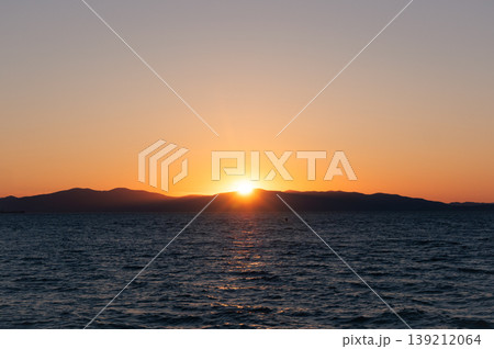 Sun dips below distant mountains painting the vast ocean with a warm, fiery glow at dusk Sun dips below distant mountains painting the vast ocean with a warm, fiery glow at dusk 139212064
