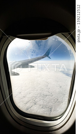 Airplane wing view from plane window over blue sky landscape in sunlight. Airplane porthole. Summer travel background. Vertical 139212512