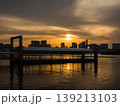 オレンジ色の夕日が海面を反射する船着き場と遠くのビルのシルエット (東京都、港区、お台場) 139213103