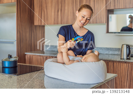 Mother talking to baby lying in cozy lounger on kitchen table while cooking at home. Caring mom interacting with infant during daily routine. Parenting, multitasking and family life concept Mother talking to baby lying in cozy lounger on kitchen table while cooking at home. Caring mom interacting with infant during daily routine. Parenting, multitasking and family life concept 139213148
