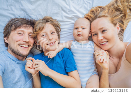 Baby lying on bed with mother, father and older brother at home. Happy family spending time together, bonding and relaxing. Parenthood, sibling connection and warm family life concept Baby lying on bed with mother, father and older brother at home. Happy family spending time together, bonding and relaxing. Parenthood, sibling connection and warm family life concept 139213158