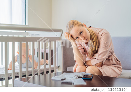 Mother sitting on sofa calculating monthly expenses with receipts while baby lies in crib nearby at home. Stressed mom managing budget and childcare. Financial pressure and parenthood concept 139213194