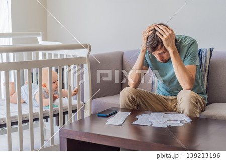 Father sitting on sofa calculating monthly expenses with receipts while baby lies in crib nearby at home. Stressed dad managing budget and childcare. Financial pressure and parenthood concept 139213196