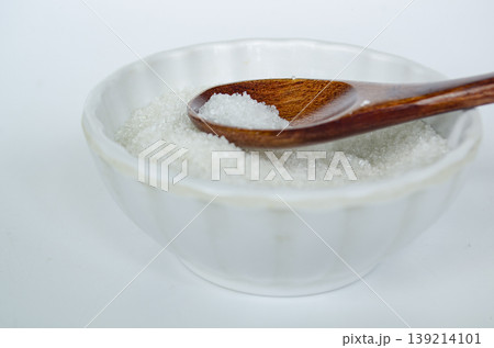 Side view of salt in wooden spoon inside white bowl highlighting seasoning ingredient and controlled sodium consumption. 139214101