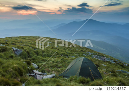 Summer camping in mountains at dawn. Tourist tent on round grassy hill on distant misty blue mountains range under pink sky before sunrise or sunset. Tourism, hiking and beauty of nature concept. Summer camping in mountains at dawn. Tourist tent on round grassy hill on distant misty blue mountains range under pink sky before sunrise or sunset. Tourism, hiking and beauty of nature concept. 139214887