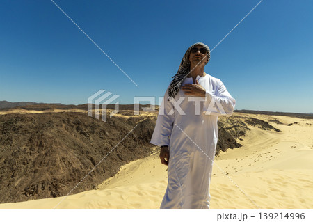Arab man in national dress stands in the Sinai or Sahara desert and looks at the blue sky. Man in traditional thobe tobe thawb dishdasha kandura and sunglasses standing proudly in desert. 139214996