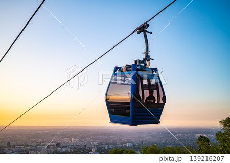 Blue cable car gondola suspended on wire against orange sunset sky Blue cable car gondola suspended on wire against orange sunset sky 139216207