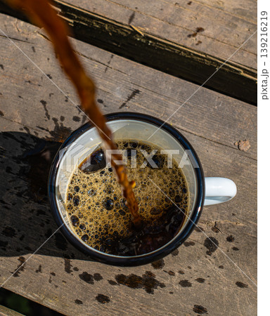 Hot coffee spilling overflowing from white enamel mug on rustic outdoor camping Hot coffee spilling overflowing from white enamel mug on rustic outdoor camping 139216219