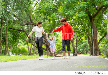 Front view Asian father, mother and daughter hold hands and walk together along the road in public park with day light, daughter look enjoy and fun with her family. Front view Asian father, mother and daughter hold hands and walk together along the road in public park with day light, daughter look enjoy and fun with her family. 139216372