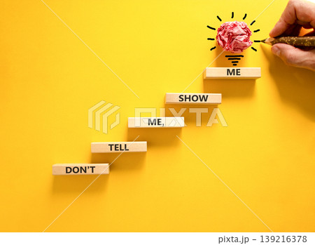 Tell or show symbol. Concept words Do not tell me, show me on beautiful wooden blocks. Beautiful yellow paper background. Light bulb icon. Businessman hand. Business tell or show concept. Copy space. 139216378