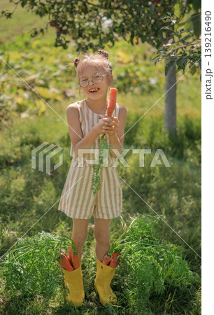 A laughing, cheerful child eats fresh carrots. A bunch of carrots sticking out of the girl's yellow rain boots A laughing, cheerful child eats fresh carrots. A bunch of carrots sticking out of the girl's yellow rain boots 139216490