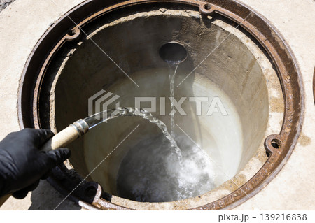 Worker in black gloves sprays water into an open manhole with a hose Worker in black gloves sprays water into an open manhole with a hose 139216838