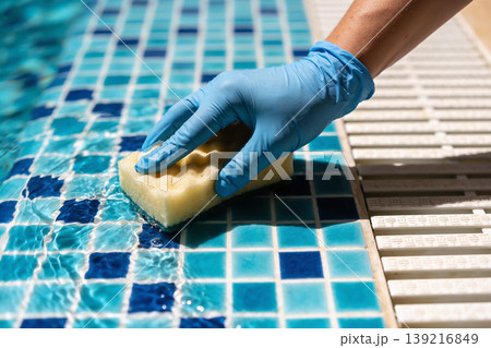 Hand in blue glove scrubbing tiled pool edge with a yellow sponge and water Hand in blue glove scrubbing tiled pool edge with a yellow sponge and water 139216849