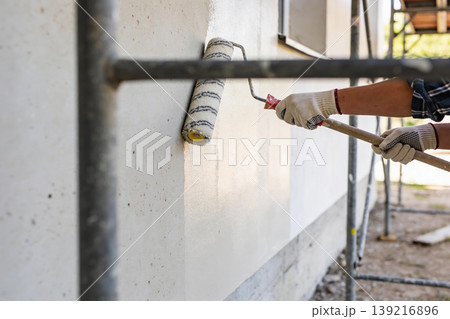 Construction worker applying white paint to exterior wall with a roller and extension pole Construction worker applying white paint to exterior wall with a roller and extension pole 139216896