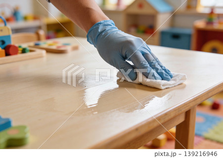 Hand in blue glove cleaning a wooden table with a white cloth in a classroom Hand in blue glove cleaning a wooden table with a white cloth in a classroom 139216946