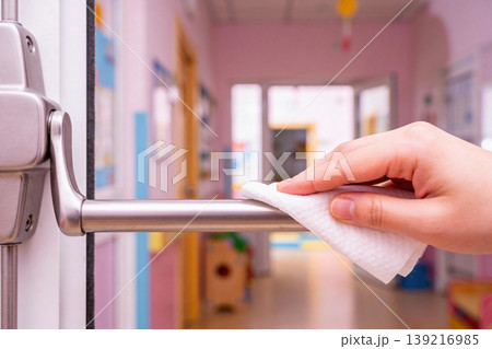 Close-up of a hand wiping a metal door handle with a white wet wipe in a brightly lit hallway 139216985