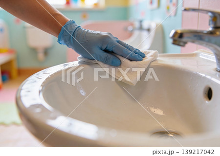 Hand wearing blue glove wipes down a white sink with a cloth for hygiene and cleanliness Hand wearing blue glove wipes down a white sink with a cloth for hygiene and cleanliness 139217042