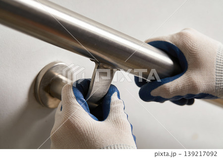 Hands in protective gloves installing a stainless steel grab bar on a white wall 139217092