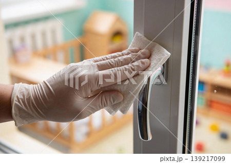 Gloved hand wipes down a window handle in a brightly lit room with toys in the background 139217099