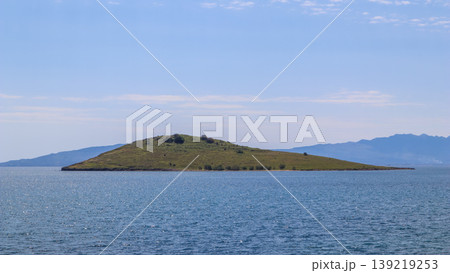 Small green island floating on tranquil blue sea, distant mountains on the horizon, bright sky with light clouds 139219253