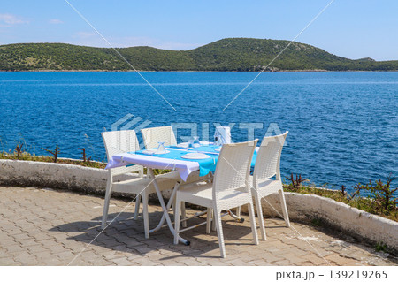 Outdoor restaurant table setting by the Aegean Sea in Bitez, Bodrum, Turkey, with calm blue water and green hills Outdoor restaurant table setting by the Aegean Sea in Bitez, Bodrum, Turkey, with calm blue water and green hills 139219265