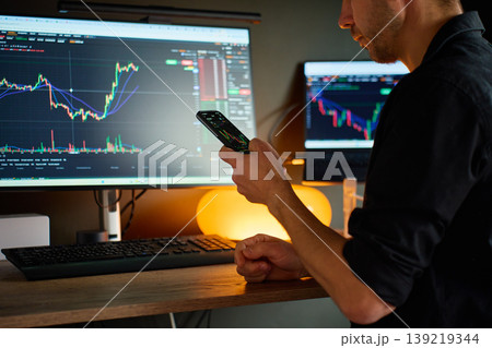 Trader checking market charts on smartphone at computer desk Trader checking market charts on smartphone at computer desk 139219344