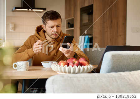 Man checking smartphone while eating breakfast at home Man checking smartphone while eating breakfast at home 139219352