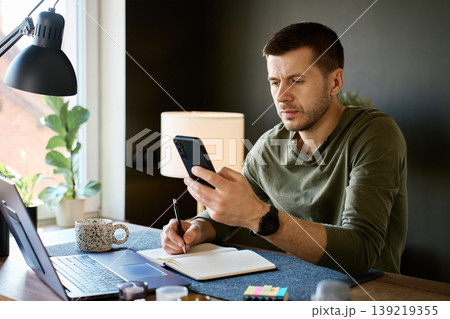 Man using smartphone and writing notes at home desk Man using smartphone and writing notes at home desk 139219355