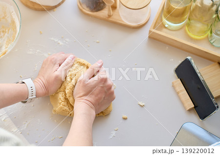Hands knead dough on a kitchen counter with flour, bowl, jars and phone on a stand Hands knead dough on a kitchen counter with flour, bowl, jars and phone on a stand 139220012