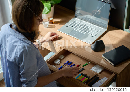 Woman arranging stationery in open desk drawer near laptop in home office 139220013