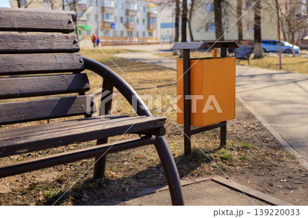 Orange metal trash bin in a city park next to a pedestrian path and a brown wooden bench. Municipal trash container designed to combat litter 139220033