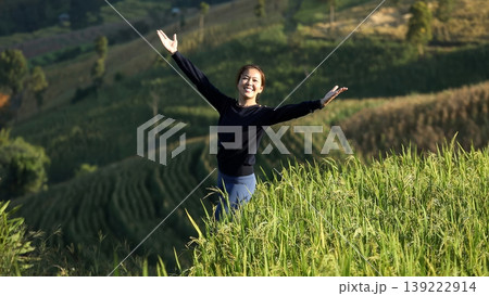 Happy Asian woman standing in green rice field on mountain, turning and raising arms with joy, celebrating nature, freedom, and healthy rural lifestyle in peaceful environment Happy Asian woman standing in green rice field on mountain, turning and raising arms with joy, celebrating nature, freedom, and healthy rural lifestyle in peaceful environment 139222914