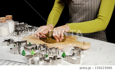 Close-up of hands kneading dark green cookie dough on wooden board, surrounded by metal Christmas cookie cutters and festive tree decorations 139223060