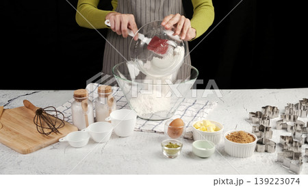 Woman pouring flour into mixing bowl, surrounded by baking ingredients and cookie cutters, preparing to bake homemade cookies 139223074