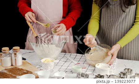 Two women baking cookies together as one whisks flour, other mixes cookie dough. Cozy kitchen scene with baking tools, matcha powder, and butter on counter 139223077