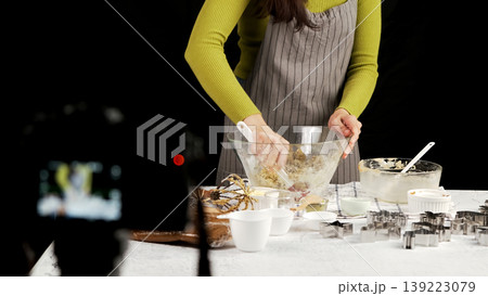 One woman in striped apron mixes cookie dough in glass bowl during baking session, filmed behind scenes with kitchen tools and cookie cutters on table One woman in striped apron mixes cookie dough in glass bowl during baking session, filmed behind scenes with kitchen tools and cookie cutters on table 139223079