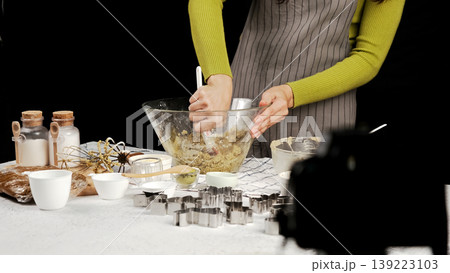 Close-up of hands mixing cookie dough in glass bowl, surrounded by baking tools and cookie cutters, with camera capturing baking process behind scenes 139223103