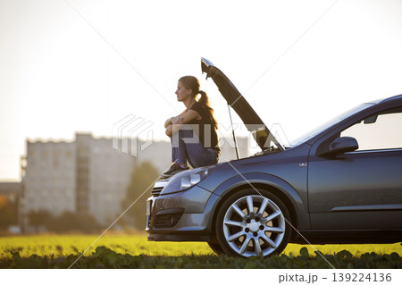 Profile of young slim attractive woman sitting on car with popped hood in green meadow waiting for help on clear sky copy space background. Transportation, vehicles problems and breakdowns concept. Profile of young slim attractive woman sitting on car with popped hood in green meadow waiting for help on clear sky copy space background. Transportation, vehicles problems and breakdowns concept. 139224136
