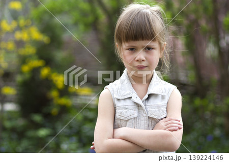 Pretty funny moody little blond preschool girl in white sleeveless dress looks into camera feeling angry and unsatisfied on blurred summer background. Children tantrum concept. Pretty funny moody little blond preschool girl in white sleeveless dress looks into camera feeling angry and unsatisfied on blurred summer background. Children tantrum concept. 139224146