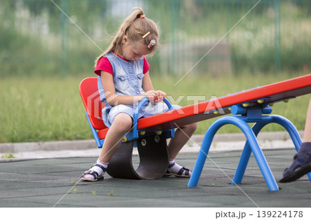 Playground conflicts. Small cute young blond child girl sits moody, angry and offended on see- saw swing on bright green blurred background. Joys and sorrows of childhood, children tantrums concept. Playground conflicts. Small cute young blond child girl sits moody, angry and offended on see- saw swing on bright green blurred background. Joys and sorrows of childhood, children tantrums concept. 139224178