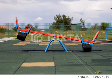 New modern plastic bright colorful blue and red empty toy seesaw swing on nursery playground with soft rubber flooring on bright sunny summer day. Perfect place for children activities outdoors. New modern plastic bright colorful blue and red empty toy seesaw swing on nursery playground with soft rubber flooring on bright sunny summer day. Perfect place for children activities outdoors. 139224219