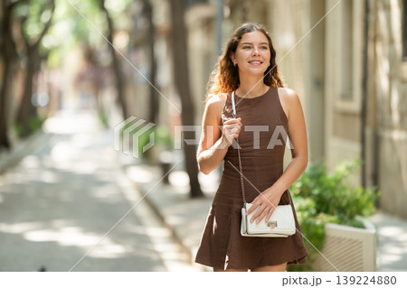 Cute young girl wearing stylish short summer dress with white handbag on shoulder walking along shady tree lined street enjoying spring day in Barcelona Cute young girl wearing stylish short summer dress with white handbag on shoulder walking along shady tree lined street enjoying spring day in Barcelona 139224880
