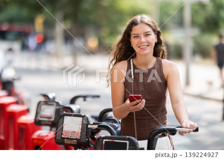 Stylish young girl renting bike using app on her phone while walking through streets of Barcelona Stylish young girl renting bike using app on her phone while walking through streets of Barcelona 139224927