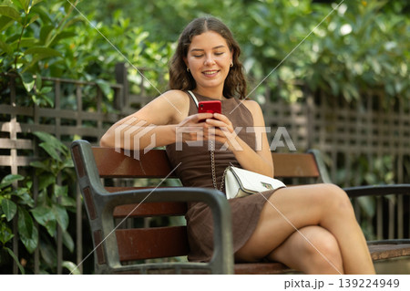 young woman sits on a bench in summer with a phone in her hands 139224949