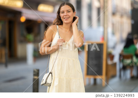 Cheerful girl in white dress walking along the street among architecture with bag 139224993
