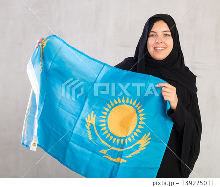 Smiling joyful Muslim woman in traditional black hijab holds flag of Kazakhstan Smiling joyful Muslim woman in traditional black hijab holds flag of Kazakhstan 139225011