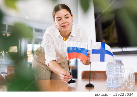 Positive young woman putting flag of Russia on the table with flag of Finland in conference room 139225034