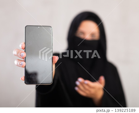 young Muslim girl with her face covered by burka shows empty dark smartphone screen. 139225189