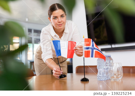 Positive young woman putting flag of France on the table with flag of Norway in conference room Positive young woman putting flag of France on the table with flag of Norway in conference room 139225234