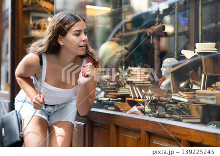 Interested girl looking at antique handicrafts in windows of souvenir shop Interested girl looking at antique handicrafts in windows of souvenir shop 139225245
